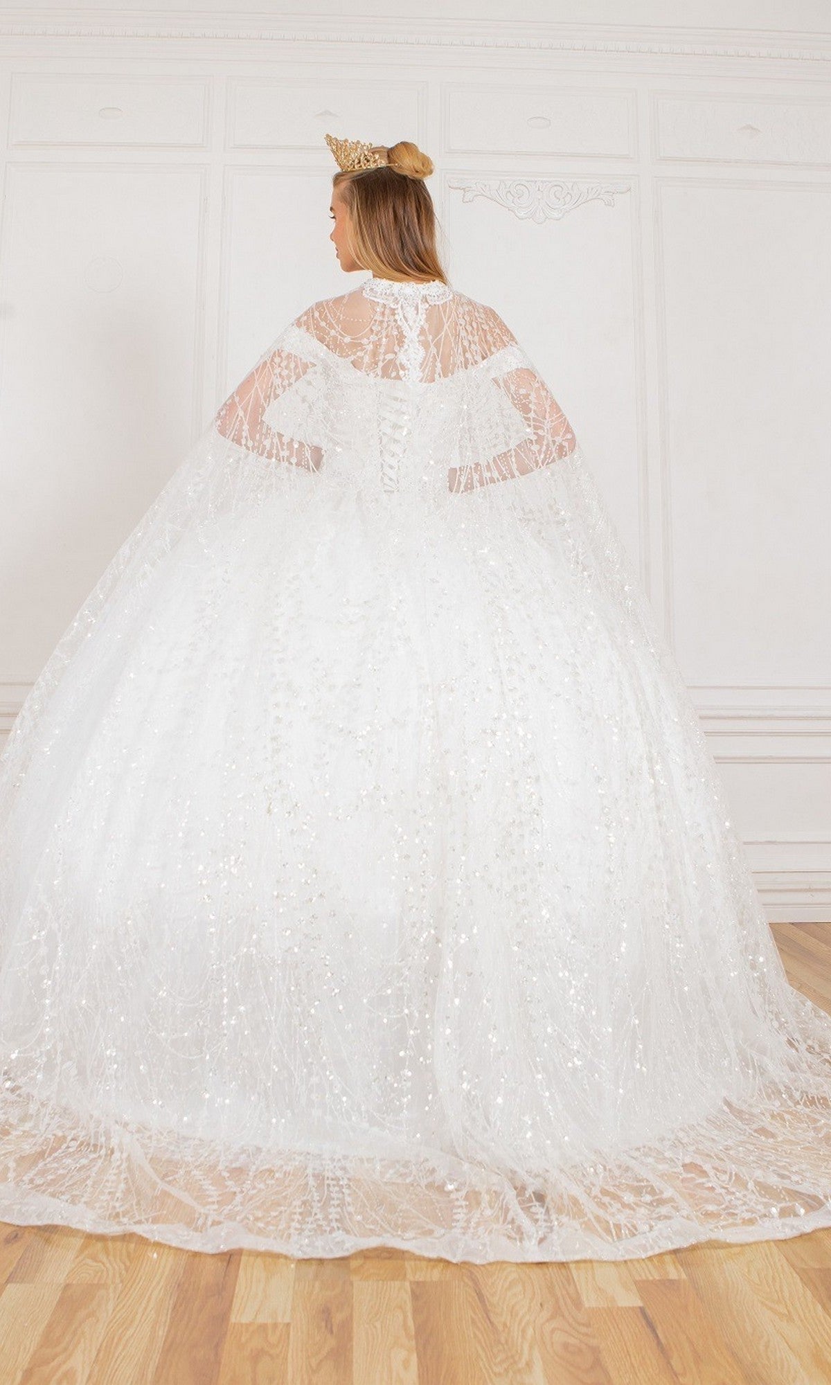 A person wearing the Quinceanera Dress 4201 by Cinderella Couture and a crown stands with their back to the camera in a bright, ornate room with wooden floors.